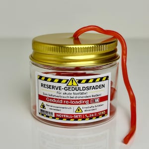 Könnte beinhalten: Ein kleines Glas mit goldenem Deckel und einer roten, lakritzartigen Süßigkeit, die aus der Oberseite herausragt. Das Glas hat ein Etikett mit deutschem Text, einschließlich "RESERVE-GEDULDSFADEN". Die Süßigkeit ist etwa 25 cm lang.