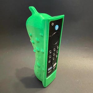 May include: A bright green, textured, pickle-shaped remote control cover housing a black television remote. The cover features a bumpy surface and a stem-like protrusion at the top, designed to fit the rectangular remote with various control buttons.