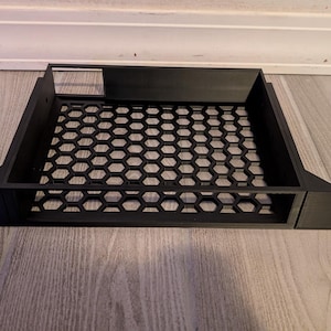 May include: Black rectangular computer component with a honeycomb pattern in the center. The component has a rectangular opening at the top and a logo on the side. The component is made of a matte black material.