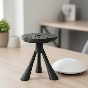 May include: A black tripod stand with a circular top featuring multiple holes. The stand has three legs and is positioned on a light-colored wooden surface. A white, round device with a blue light is next to the stand.