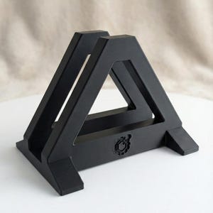 May include: A black, geometric stand with a triangular design. The stand features a solid base with two supporting feet and a decorative emblem. The structure is made of a matte black material and is designed to hold an item.