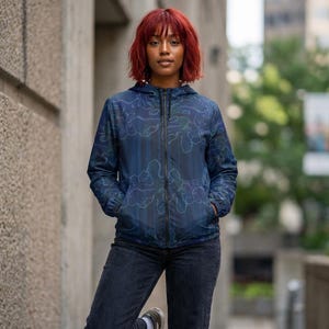 May include: A hooded, zip-up jacket in shades of blue with a pattern of white and teal cloud-like shapes. The jacket has a black zipper and pockets. The person is wearing dark wash jeans and sneakers.
