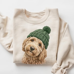 Puede incluir: Sudadera color crema con una ilustración detallada de un perro golden doodle con un gorro de punto verde con un pompón. El pelaje del perro es de color marrón claro y crema. La sudadera tiene mangas largas.