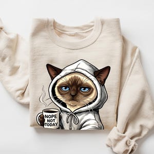 May include: Beige sweatshirt featuring a grumpy cartoon cat wearing a white hoodie and holding a coffee mug. The mug reads "Nope Not Today." The cat has blue eyes and brown ears. The sweatshirt has long sleeves.