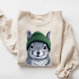 Puede incluir: Una sudadera de color crema con una ilustración detallada de una ardilla con un gorro verde. La ardilla tiene pelaje gris y una expresión sonriente. La sudadera tiene mangas largas.