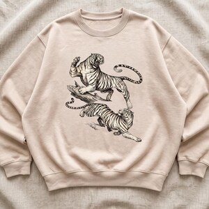 Può includere: Una felpa girocollo color beige con un'illustrazione dettagliata a inchiostro nero di due tigri. Una tigre è posizionata su un pezzo di legno con la bocca aperta, mentre la seconda tigre è raffigurata in una posa dinamica di corsa sottostante.