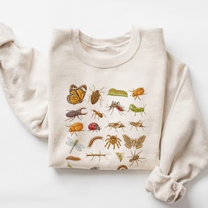 Puede incluir: Sudadera beige con una ilustración colorida de varios insectos, incluyendo una mariposa, una mariquita y una libélula. El gráfico está detallado con una variedad de colores y formas. La parte superior de manga larga está hecha de un material suave y está doblada sobre una superficie blanca.