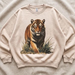 May include: A beige crewneck sweatshirt featuring a detailed graphic print of a tiger walking through tall grass. The tiger is depicted in shades of orange, black, and white, with a focused expression. The sweatshirt has long sleeves and ribbed cuffs.