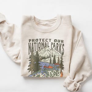 Op de afbeelding: Een beige sweatshirt met de tekst "PROTECT OUR NATIONAL PARKS" boven een afbeelding van bergen, bomen en een rivier. De sweatshirt heeft opgerolde mouwen en een ronde hals.