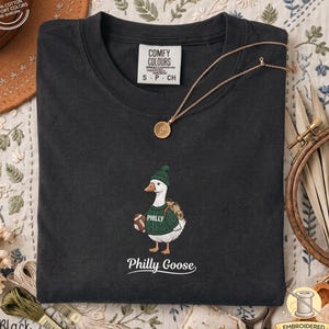 Può includere: Una t-shirt nera con il ricamo di un'oca bianca che indossa un berretto di lana verde e un maglione verde con la scritta PHILLY. L'oca tiene in mano un pallone da football e porta uno zaino. Il testo Philly Goose si trova in basso.
