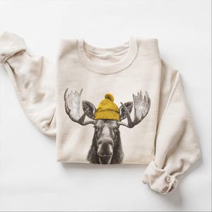 May include: A cream-colored sweatshirt featuring a detailed illustration of a moose wearing a yellow knit beanie with a pom-pom. The moose has large antlers and a dark brown face. The sleeves are rolled up.