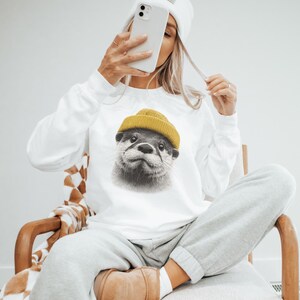 Op de afbeelding: Wit sweatshirt met lange mouwen met een gedetailleerde illustratie van een otter met een gele beanie. Het gezicht van de otter is het middelpunt, met een neutrale uitdrukking. Het sweatshirt wordt gecombineerd met grijze joggingbroeken.