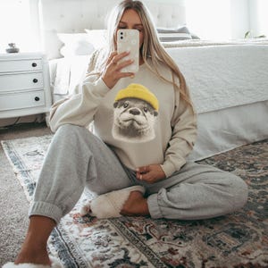 Op de afbeelding: Beige sweatshirt met een gedetailleerde otter met een gele beanie. De sweatshirt wordt gecombineerd met grijze joggingbroek. De otter-afbeelding is gecentreerd op de voorkant. De persoon zit op een patroonkleed.