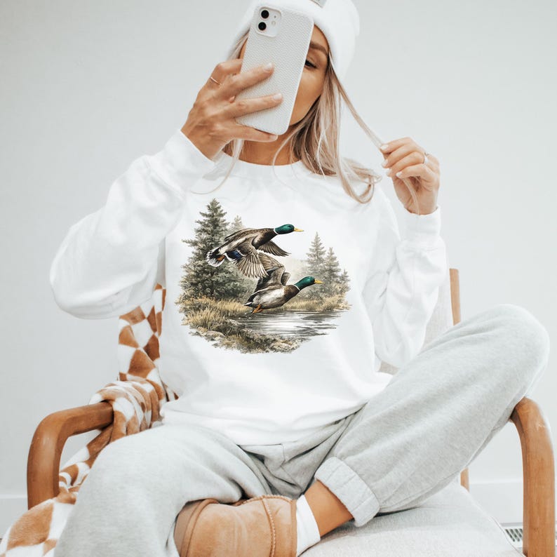 K&ouml;nnte beinhalten: Wei&szlig;es Langarm-Sweatshirt mit einer Naturszene, die zwei fliegende Enten &uuml;ber Wasser zeigt, umgeben von B&auml;umen. Die Enten haben gr&uuml;ne K&ouml;pfe und braune K&ouml;rper. Das Sweatshirt ist aus einem weichen Material gefertigt.