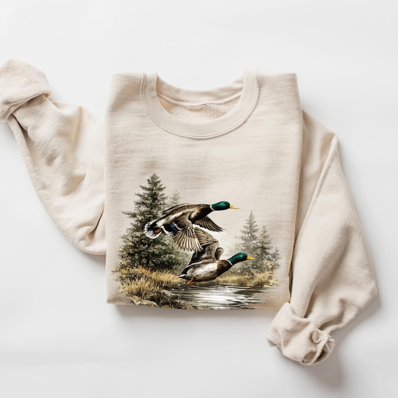 K&ouml;nnte beinhalten: Beiges Sweatshirt mit detaillierter Grafik von drei Enten im Flug &uuml;ber Wasser, mit immergr&uuml;nen B&auml;umen im Hintergrund. Die Enten haben gr&uuml;ne K&ouml;pfe, braune K&ouml;rper und gelbe Schn&auml;bel. Das Sweatshirt hat lange &Auml;rmel.