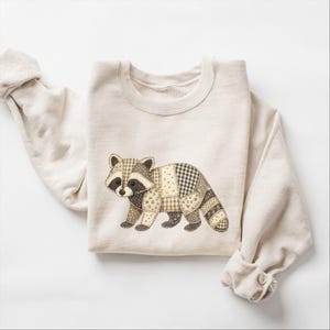May include: A cream-colored crewneck sweatshirt featuring a central graphic of a raccoon. The raccoon is designed with a patchwork aesthetic, utilizing various brown, beige, and black fabric patterns including gingham, polka dots, and floral prints.