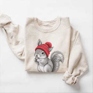 Puede incluir: Una sudadera de cuello redondo color crema con una ilustración detallada de una ardilla gris que lleva un gorro de punto rojo brillante con un pompón. La ardilla aparece sentada, sosteniendo una pequeña bellota marrón entre sus patas delanteras.
