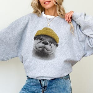 Op de afbeelding: Een lichtgrijs sweatshirt met een afbeelding van een otter met een mosterdgele beanie. Het gezicht van de otter is in zwart en wit. De sweatshirt heeft een ronde hals en lange mouwen.