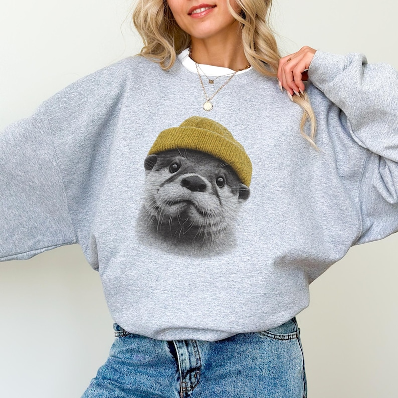 Op de afbeelding: Een lichtgrijs sweatshirt met een afbeelding van een otter met een mosterdgele beanie. Het gezicht van de otter is in zwart en wit. De sweatshirt heeft een ronde hals en lange mouwen.