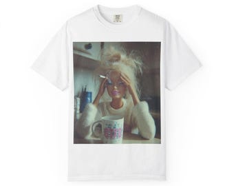 May include: White t-shirt featuring a printed image of a Barbie doll with messy blonde hair, smoking a cigarette, and holding her head. A mug with the text "GET FED" is in front of her.