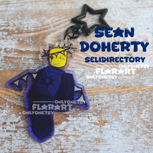 Puede incluir: Un llavero acrílico transparente con un personaje de dibujos animados con una cara amarilla, una corona y una capa azul oscuro. El llavero tiene un dije en forma de estrella negra. El texto "SEAN DOHERTY" y "FLAR ART" son visibles.