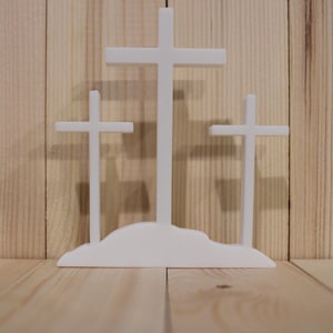 May include: A white, three-cross religious display. The crosses are of varying heights, with the tallest in the center. The crosses are mounted on a white base, set against a light wood background.