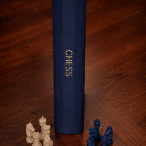 May include: A navy blue book with the word "CHESS" in gold lettering. The book is standing upright next to chess pieces in cream and navy blue. The text "Playbook'd Chess" is in the upper left corner.