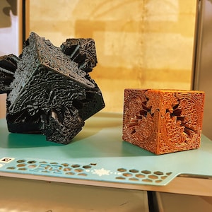 May include: Two intricately designed, geometric sculptures. One is a dark, complex cube with protruding sections. The other is a smaller, orange-brown cube with a carved, ornamental pattern. Both are on a light blue surface.