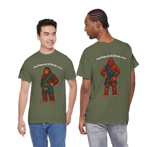 Puede incluir: Dos camisetas verde oliva con un dibujo animado de Bigfoot con un kilt escocés. La parte delantera de las camisetas muestra el texto "TheSquatchShop.com" encima del gráfico de Bigfoot. La parte trasera de las camisetas también muestra el texto "TheSquatchShop.com".