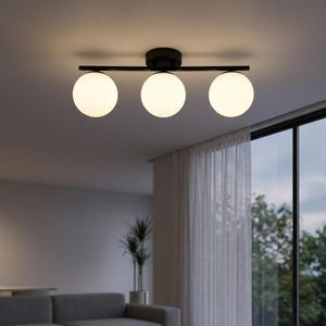 Puede incluir: Un moderno plafón con tres pantallas esféricas de vidrio esmerilado. Las pantallas están dispuestas a lo largo de una barra negra. La lámpara está montada en un techo oscuro, iluminando la habitación.