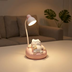 Puede incluir: Una lámpara de escritorio caprichosa con una figura de gato blanco con antifaz, sentada en un cojín rosa. La lámpara tiene un cuello flexible y una pantalla blanca con la parte superior rosa. El gato sostiene un libro, creando un ambiente relajante.