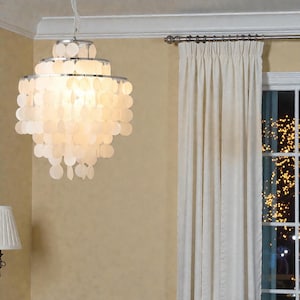 May include: A decorative chandelier with multiple tiers, featuring a chrome frame and white, shell-shaped elements. The chandelier hangs in a room with a beige wall, a cream curtain, and a window. The light fixture is suspended from the ceiling.