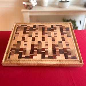Pu&ograve; includere: Tagliere rettangolare in legno con motivo a mosaico. &Egrave; realizzato con una miscela di blocchi di legno chiari e scuri, creando un design accattivante. I bordi sono incorniciati con legno pi&ugrave; chiaro e una scanalatura per il succo corre lungo il perimetro. Misura circa 30 cm x 30 cm.