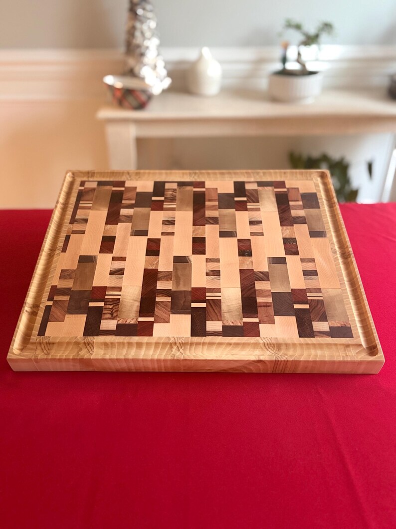 Pu&ograve; includere: Tagliere rettangolare in legno con motivo a mosaico. &Egrave; realizzato con una miscela di blocchi di legno chiari e scuri, creando un design accattivante. I bordi sono incorniciati con legno pi&ugrave; chiaro e una scanalatura per il succo corre lungo il perimetro. Misura circa 30 cm x 30 cm.