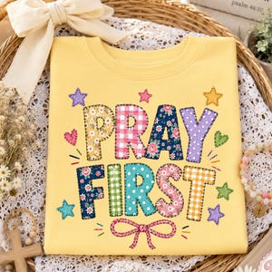 Könnte beinhalten: Ein blassgelbes Sweatshirt mit Rundhalsausschnitt und dem Schriftzug PRAY FIRST in großen, mehrfarbigen Blockbuchstaben mit verschiedenen Mustern wie Blumen, Vichy-Karo und Punkten. Das Design ist mit kleinen bunten Sternen, Herzen und einer Schleife verziert.