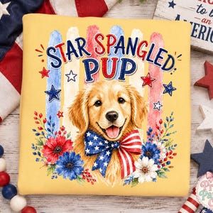 Puede incluir: Una sudadera amarilla con el gráfico de un cachorro golden retriever que lleva una pajarita con estampado de estrellas rojas, blancas y azules. El texto Star Spangled Pup está rodeado de flores y estrellas rojas y azules.