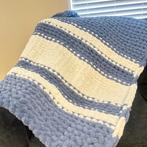 May include: A soft, chunky knit blanket in shades of blue and cream. The blanket features diagonal stripes of light blue and cream, creating a cozy and textured appearance. The blanket is folded over a dark gray surface.