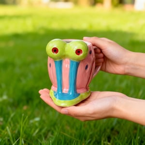 Può includere: Una tazza in ceramica a forma del personaggio Gary la lumaca di Spongebob Squarepants. La tazza è rosa con occhi verdi, strisce blu e una base verde e blu. La tazza è tenuta in due mani su uno sfondo verde sfocato.