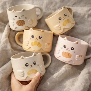 Può includere: Cinque tazze in ceramica a forma di gatto in vari colori pastello: crema, giallo e rosa. Ogni tazza presenta un design a forma di gatto e un alimento diverso sul davanti, tra cui un croissant e pane.