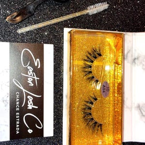 May include: False eyelashes, applicator, and lash brush on a glittery black surface. The eyelashes are in a gold case with the text "3D A04". A business card reads "Easton Lash Co. Chance Estrada". The word "Melody" is also visible.