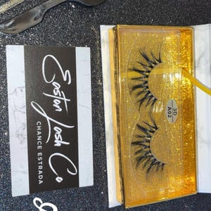 May include: A set of false eyelashes with applicators and a brush. The eyelashes are in a gold glitter box with the text "3D A02". The box is next to a business card that says "Easton Lash Co. Chance Estrada" and the name "Easlynn".