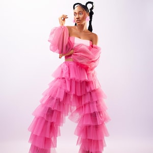 May include: A vibrant pink outfit featuring a strapless top, layered ruffle pants, and sheer, voluminous sleeves. The ensemble is accessorized with pink earrings. The model is wearing green shoes. The outfit is set against a soft, white background.
