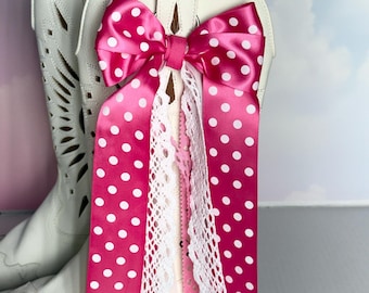 Op de afbeelding: Witte cowboylaarzen met een grote roze satijnen strik met witte stippen. De strik wordt geaccentueerd met wit kant en roze linten. De laarzen hebben decoratieve uitsparingen. De achtergrond is zacht en wazig.
