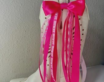 Op de afbeelding: Crèmekleurige cowboylaars met een felroze strik en bijpassende linten. De linten hebben de tekst "HOWDY" erop. De laars heeft decoratieve stiksels en uitgesneden details.