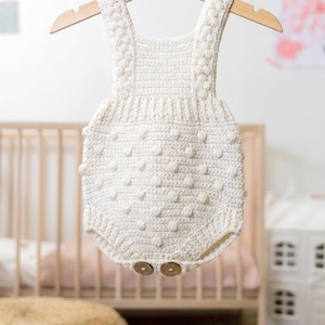 Puede incluir: Un pelele de bebé de ganchillo hecho a mano, de color crema, con un diseño de punto de burbuja texturizado. El pelele tiene botones de madera y se muestra en una percha de madera. Prenda para bebés.