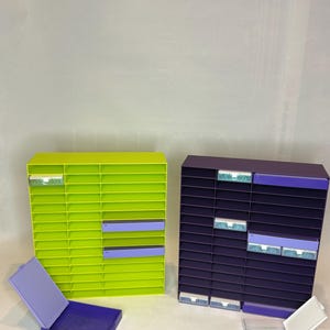 May include: Two colorful storage organizers. One is lime green with lavender accents, the other is dark purple with light purple accents. Each has multiple horizontal slots. A lavender box with a lid is open in front of the green organizer.