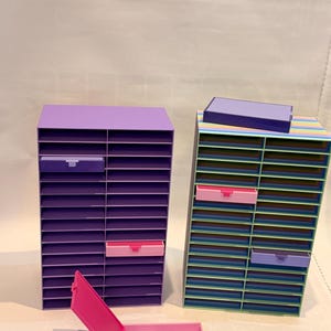May include: Two colorful desktop organizers. One is solid purple, the other is striped with blue, green, and yellow. Each has multiple slots and pull-out drawers in shades of pink and purple. A pink drawer is open in the foreground.