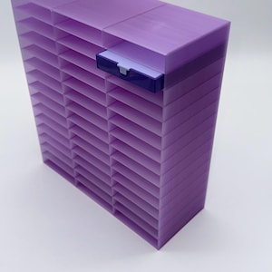May include: A rectangular, purple organizer with multiple horizontal slots. A small, dark purple drawer with a white interior is on the top right. The organizer is made of a solid, opaque material and sits on a white surface.