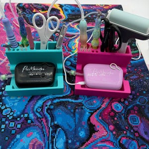 May include: Two rectangular organizers in teal and pink, each holding diamond painting tools including glittery pens, white and black scissors, and a roller. Each organizer features a small, branded electronic device labeled PicMondo.