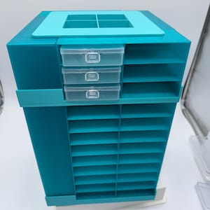 May include: A teal plastic storage organizer featuring a rotating tower design with numerous open shelves, three clear pull-out drawers, and a top section with four square compartments for sorting small items and craft supplies.